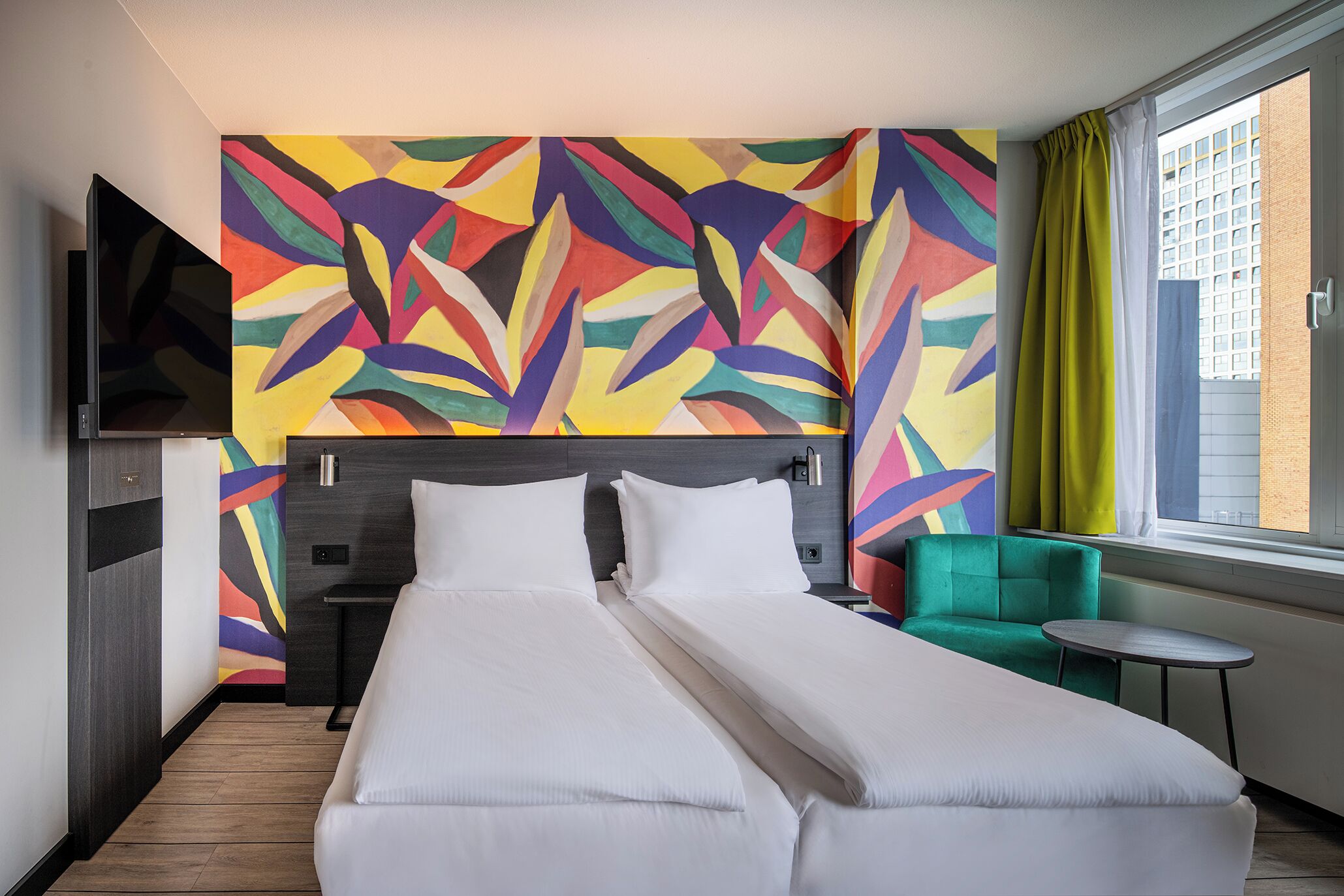 Habitación de hotel con una cama grande, un sillón verde, una mesita pequeña y un cuadro abstracto de colores en la pared.