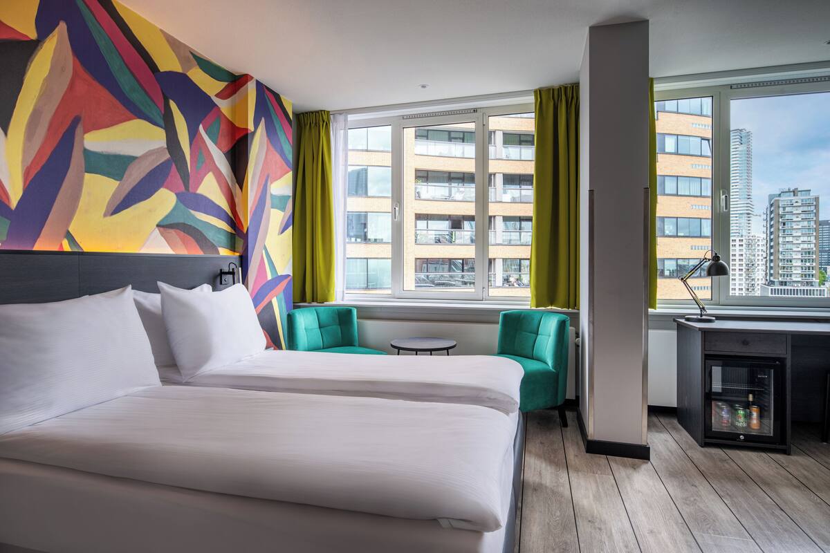 Een hotelkamer met een groot bed, twee groene fauteuils, een klein rond tafeltje en een bureau met een lamp.