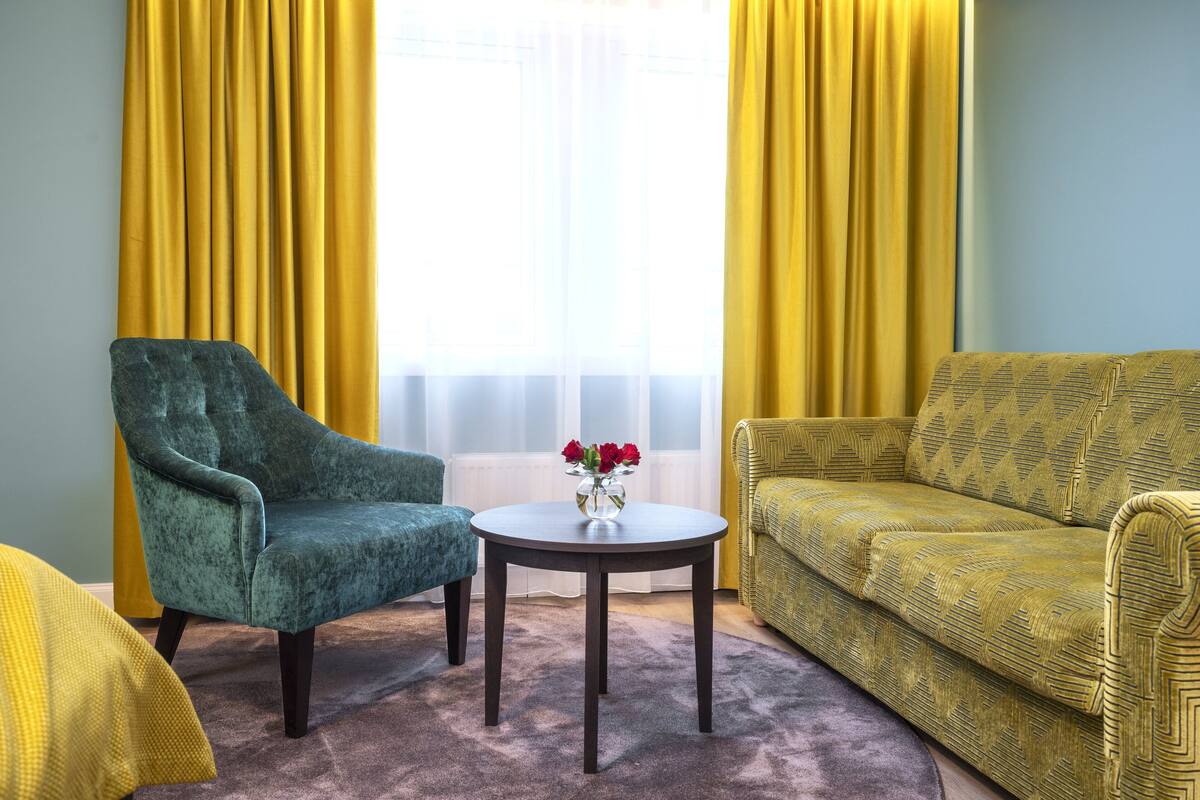 Et værelse med en turkis lænestol, en gul sofa og et rundt bord med en vase røde blomster.