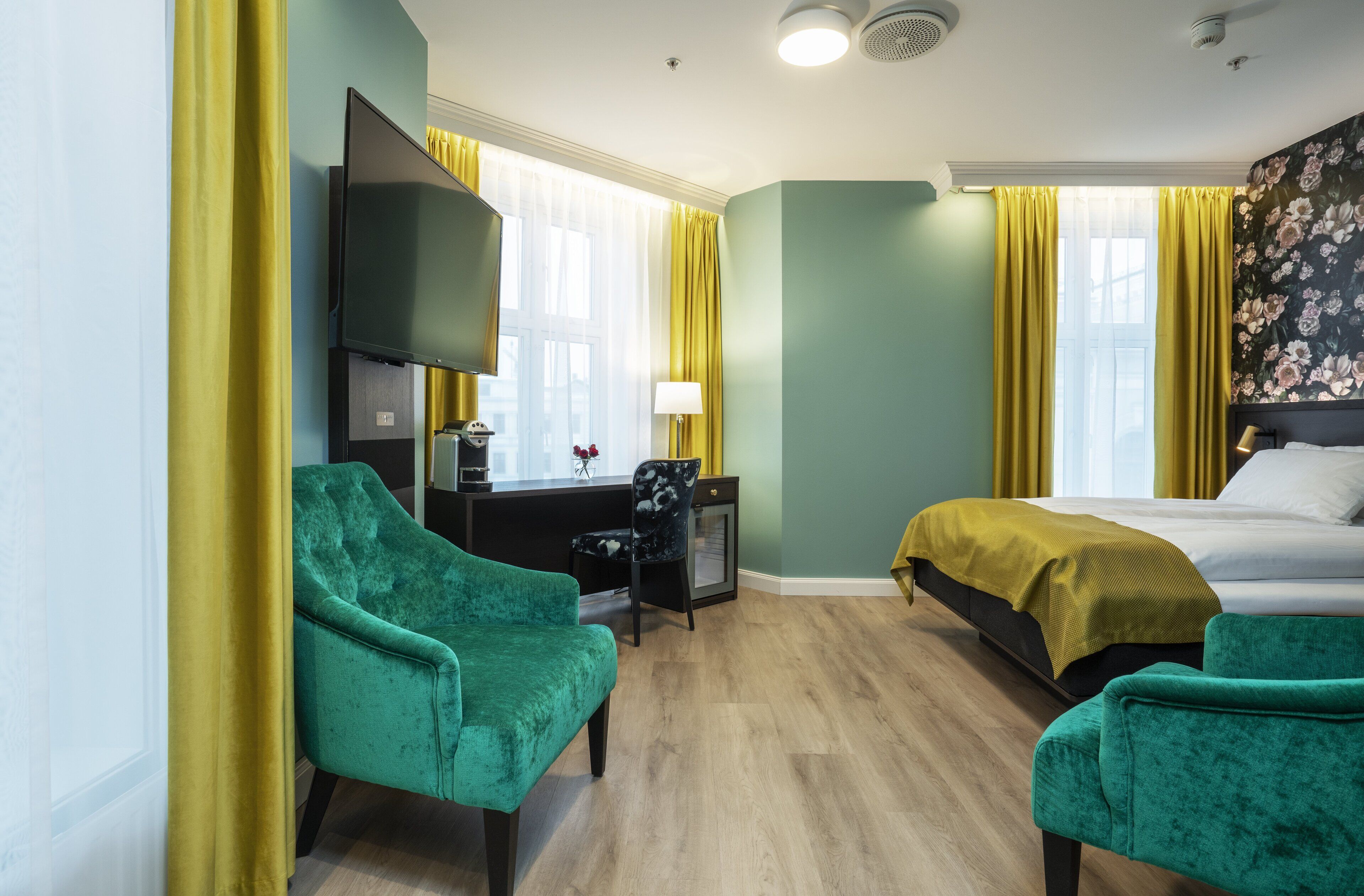 A hotel room with a bed, two green armchairs, a desk with a lamp, and a television.