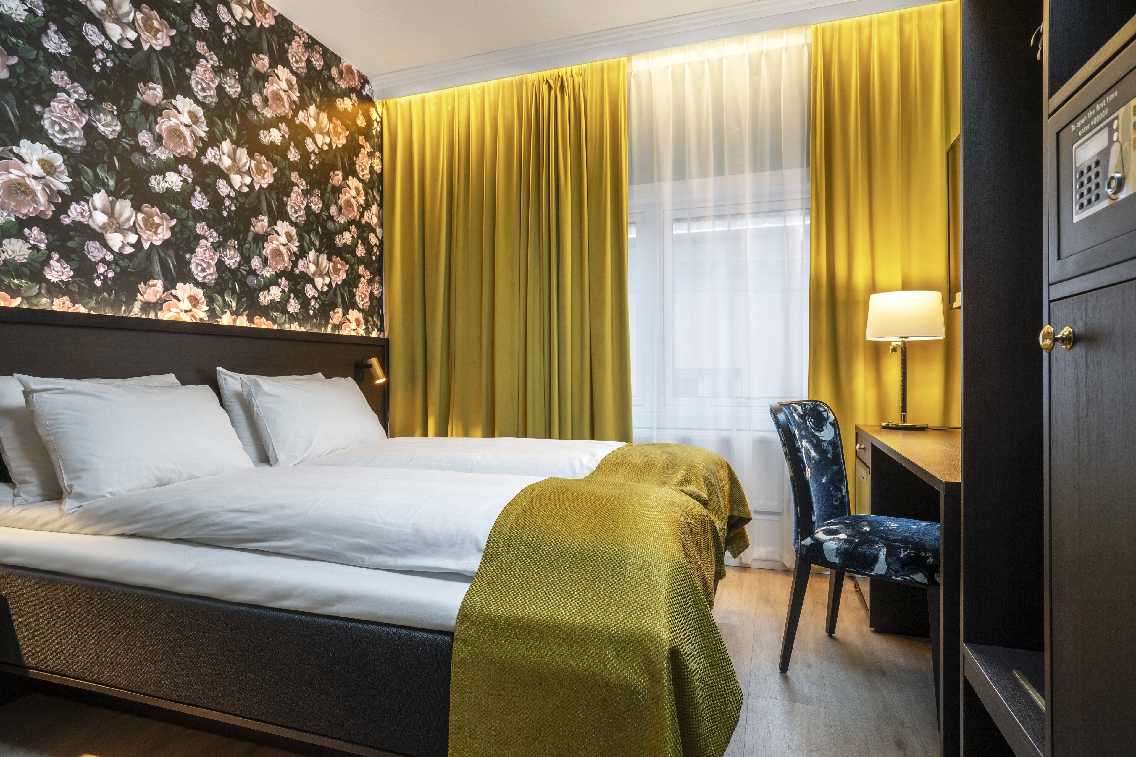 A hotel room with a bed, a desk, a chair, a lamp, and a wall with a floral pattern.