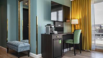 A compact hotel room with a dark wood desk, a green upholstered chair, a small refrigerator, a coffee maker, and a flat-screen TV.