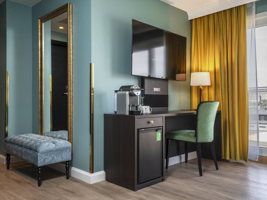 A compact hotel room with a dark wood desk, a green upholstered chair, a small refrigerator, a coffee maker, and a flat-screen TV.