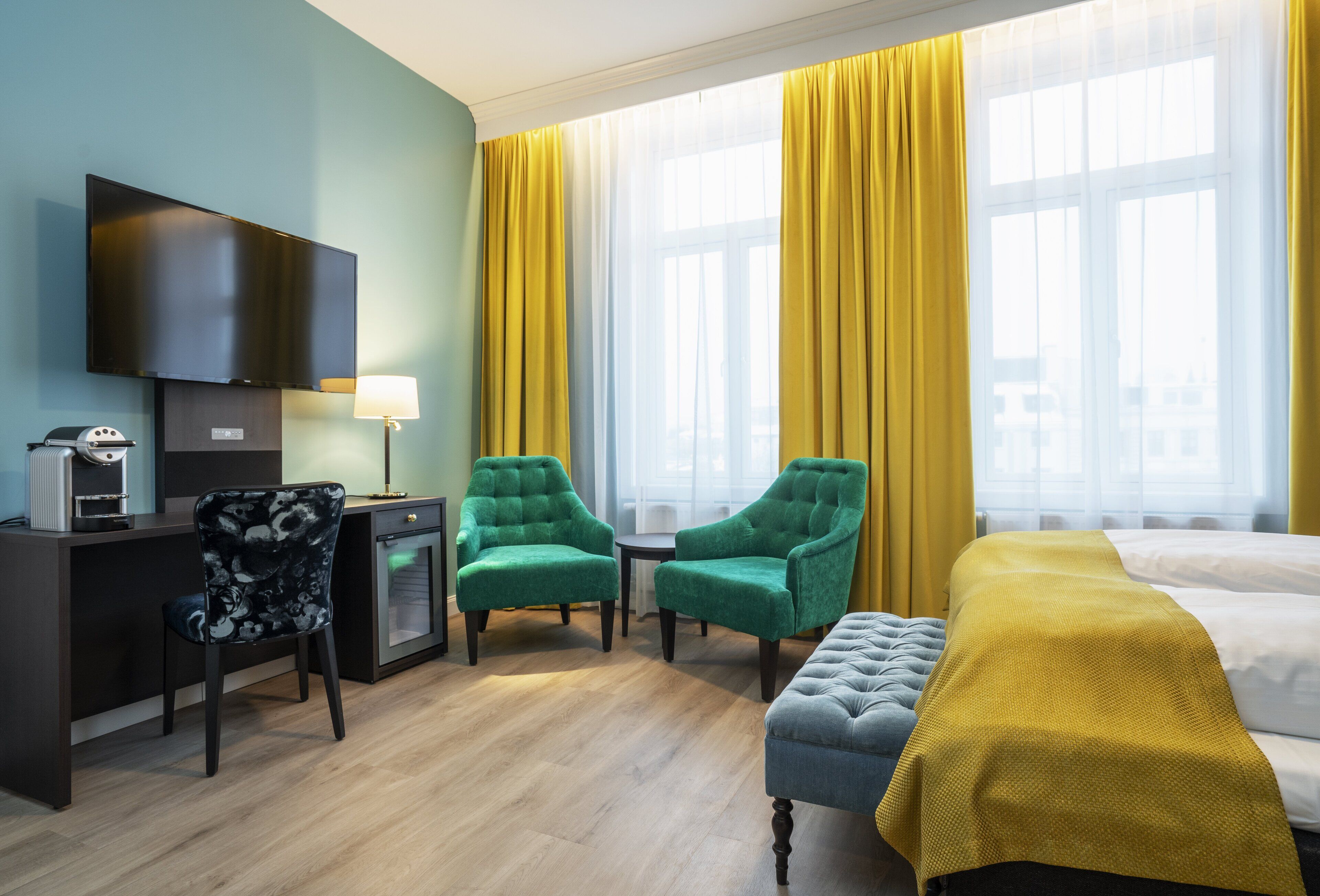 A hotel room with a desk, two green armchairs, a small table, a bed with a yellow blanket, and large windows with yellow curtains.