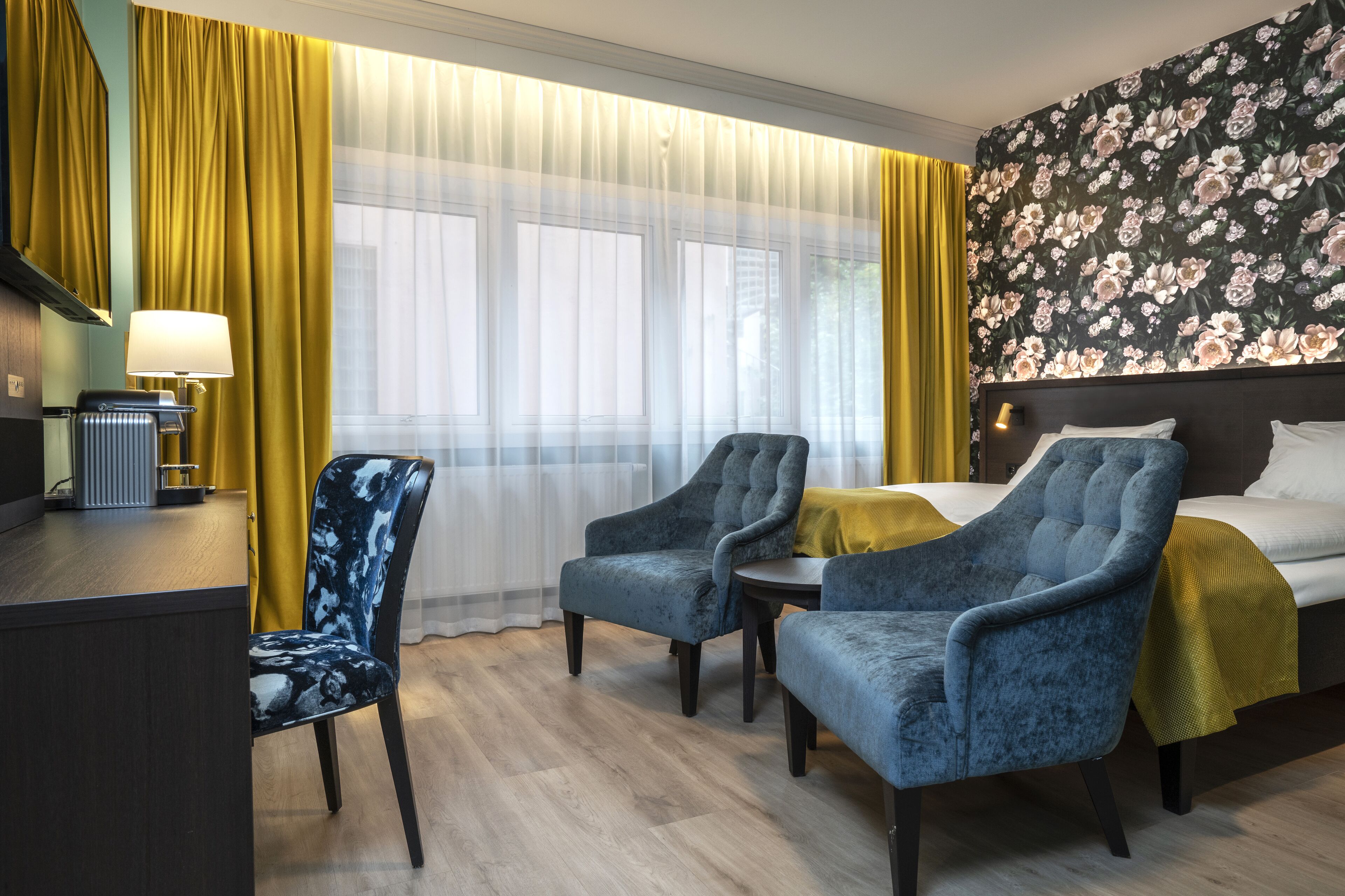 Habitación de hotel con cama, dos sillones, una mesita, un escritorio con lámpara y una ventana con cortinas.