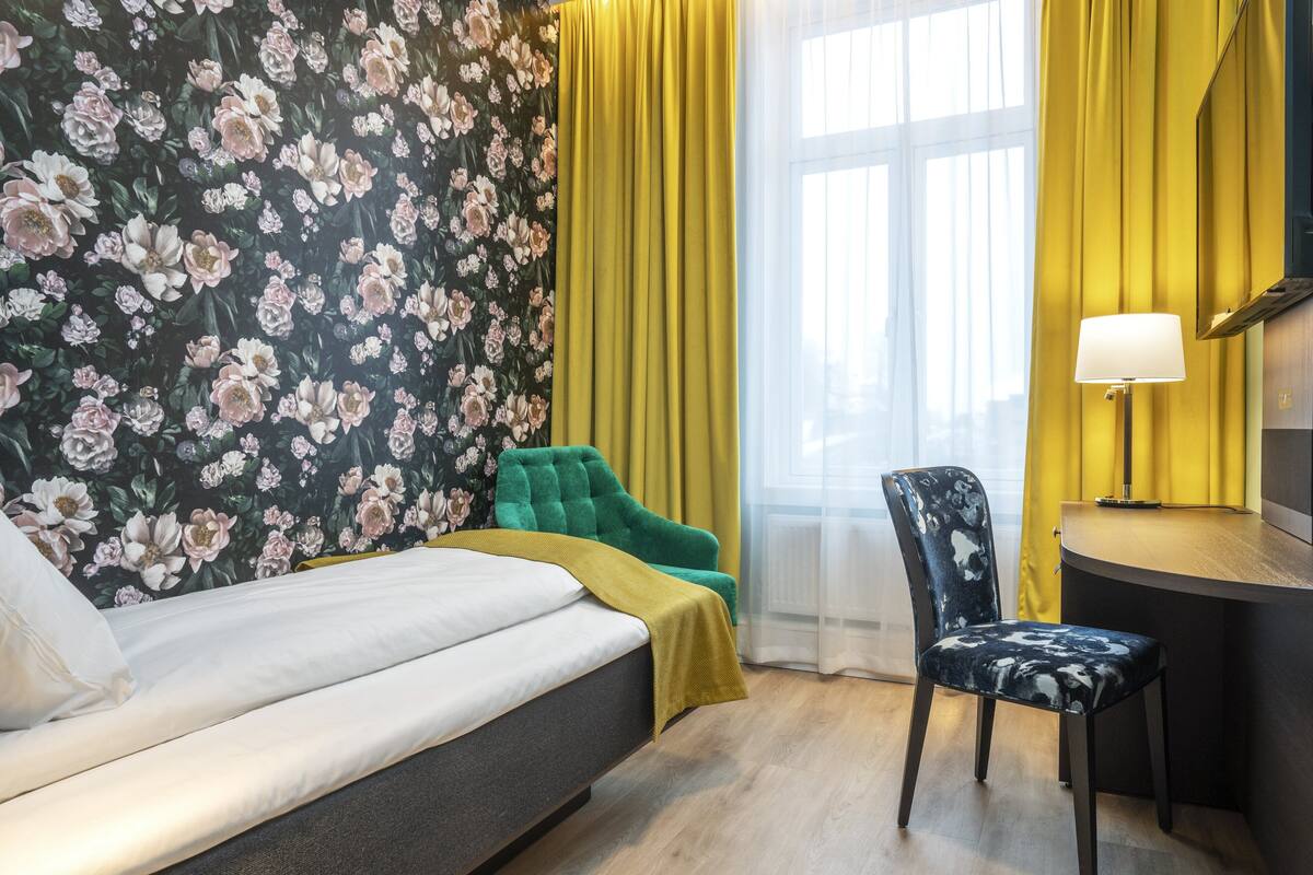 Et soveværelse med en seng, en grøn stol, et skrivebord med en lampe og tapet med blomster.
