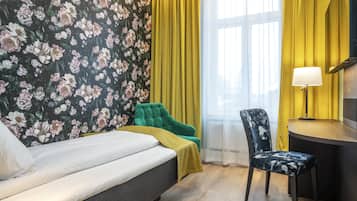 A bedroom with a bed, a green chair, a desk with a lamp, and a floral wallpaper.