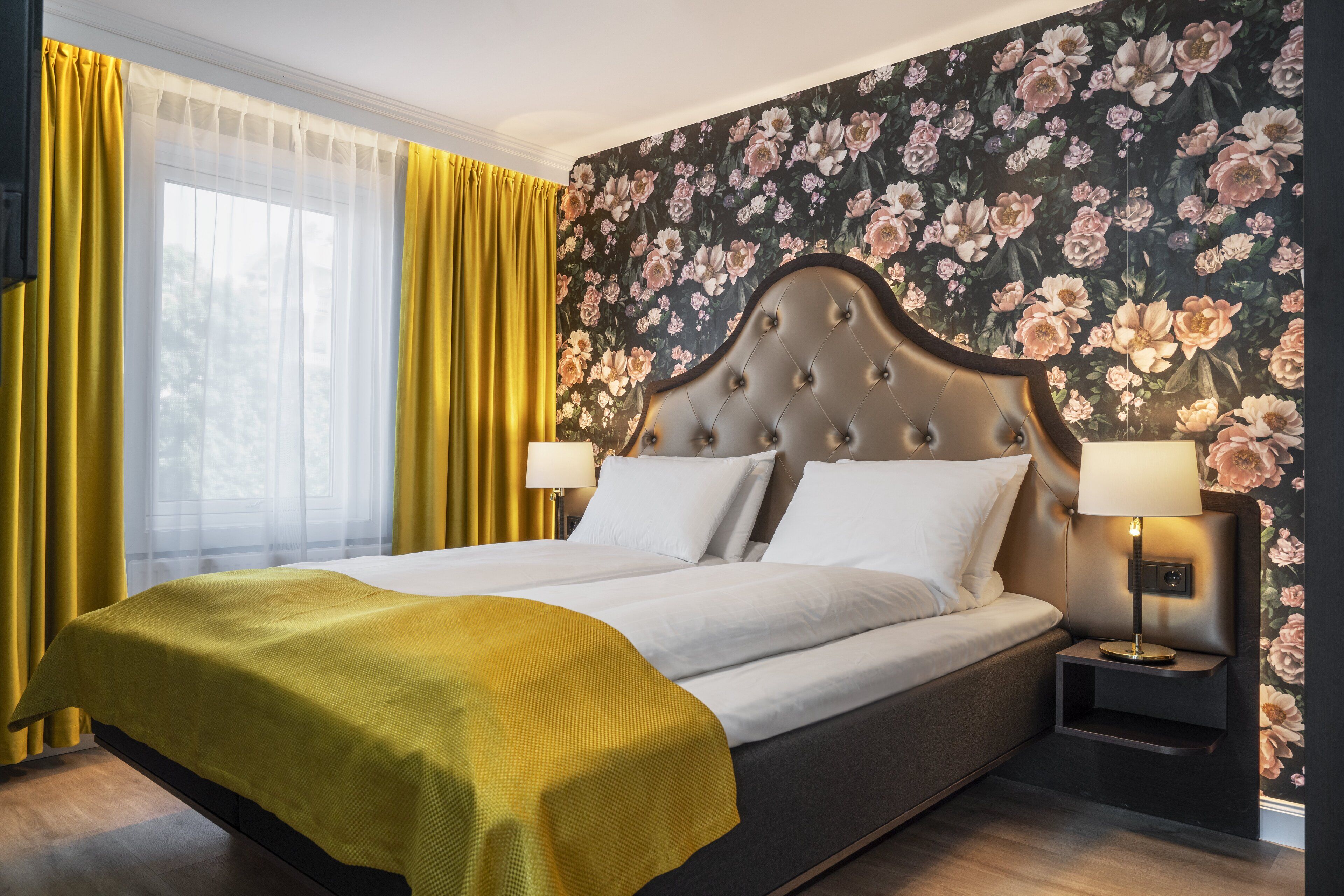 A neatly made bed with white linens, a yellow throw blanket, and a floral wallpaper backdrop.