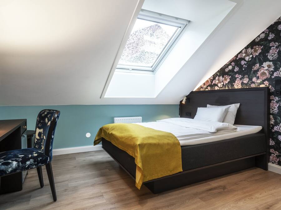 A bedroom with a skylight, a bed with a yellow blanket, a desk with a chair, and floral wallpaper.