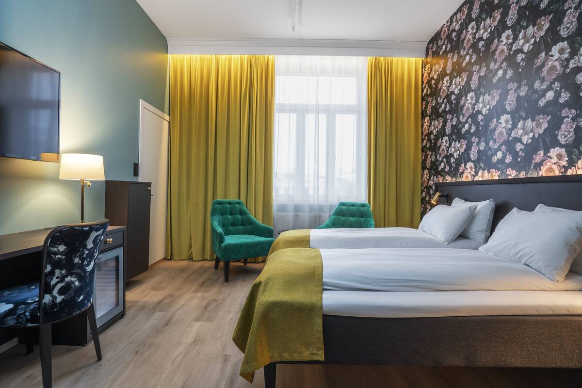 Et hotelværelse med en seng, to stole, et skrivebord, et fjernsyn og et vindue med gardiner.