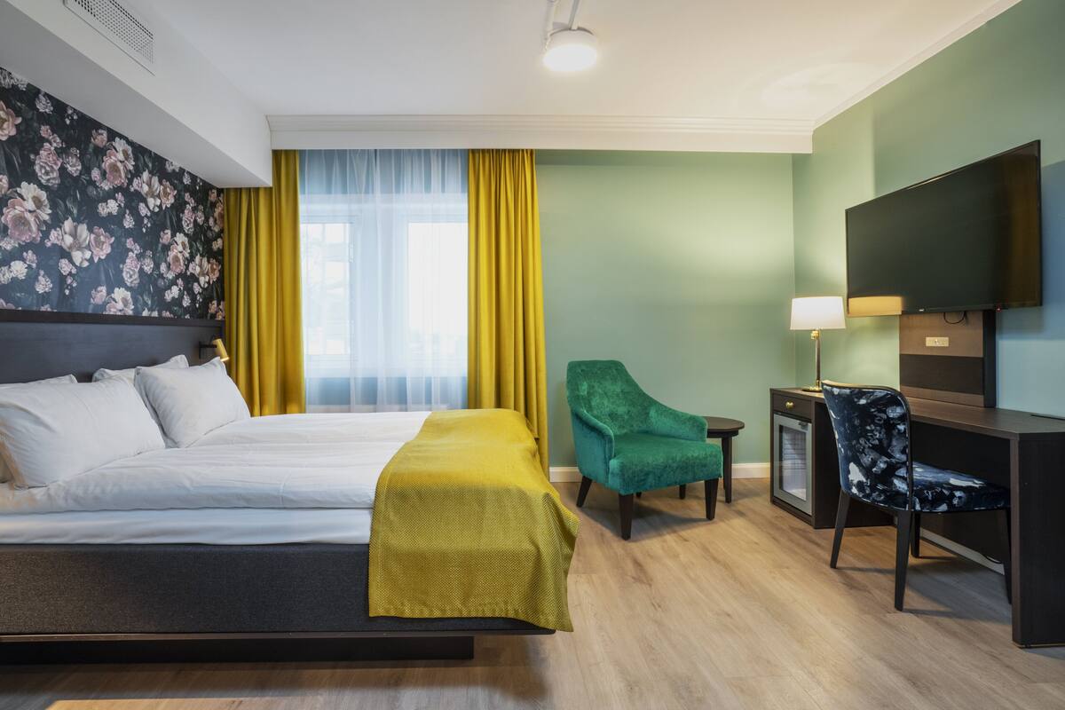 Et hotelværelse med seng, skrivebord, stol, fjernsyn og et vindue med gule gardiner.