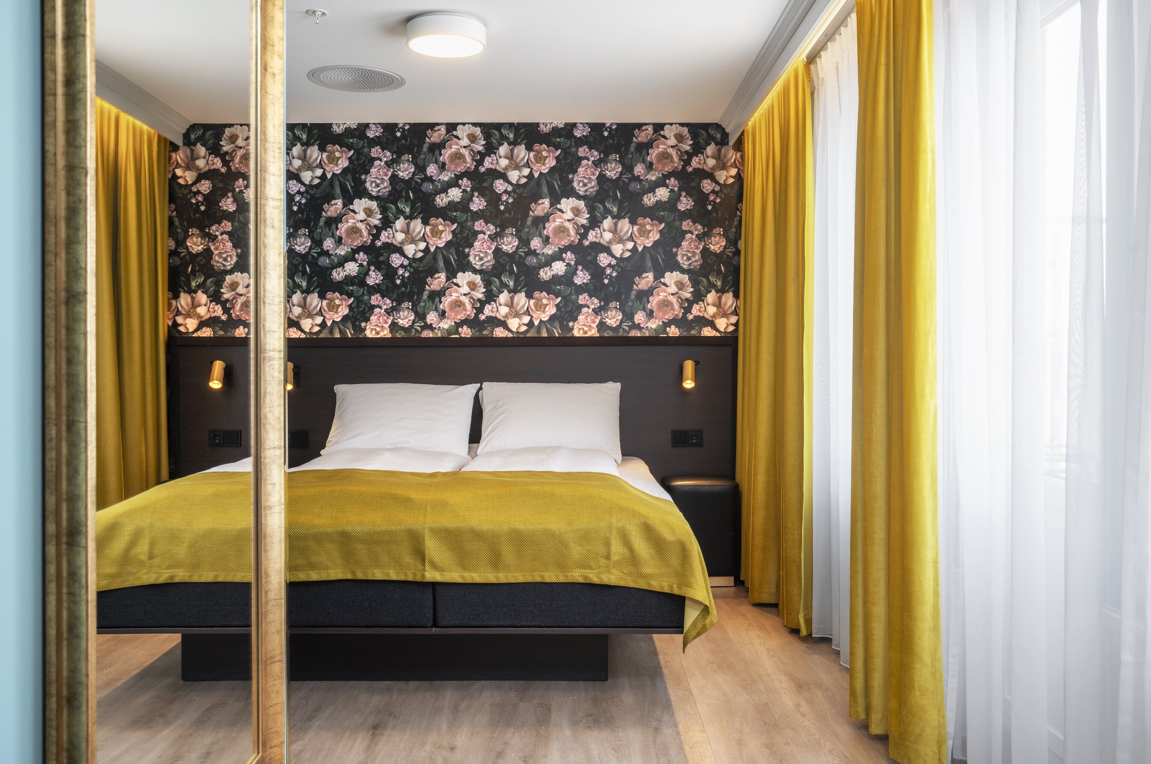 Un dormitorio moderno con una cama grande, papel tapiz floral y cortinas amarillas.