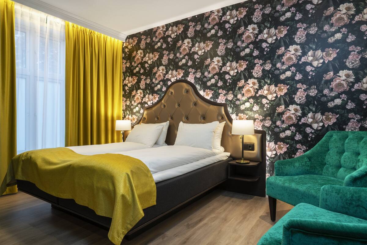 Et hotelværelse med en stor seng, en grøn lænestol, et natbord med lampe og blomstret tapet.