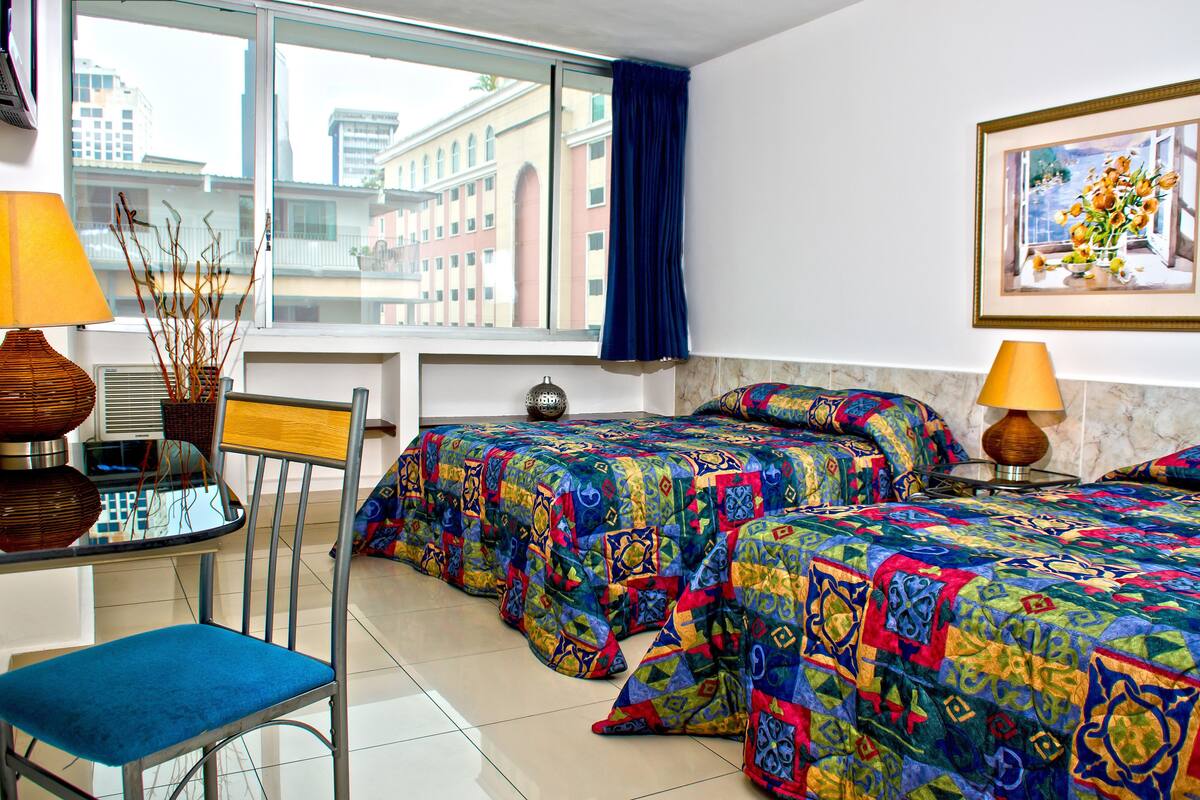 Habitación de hotel con dos camas, una silla, una mesa y un ventanal con vistas a edificios.