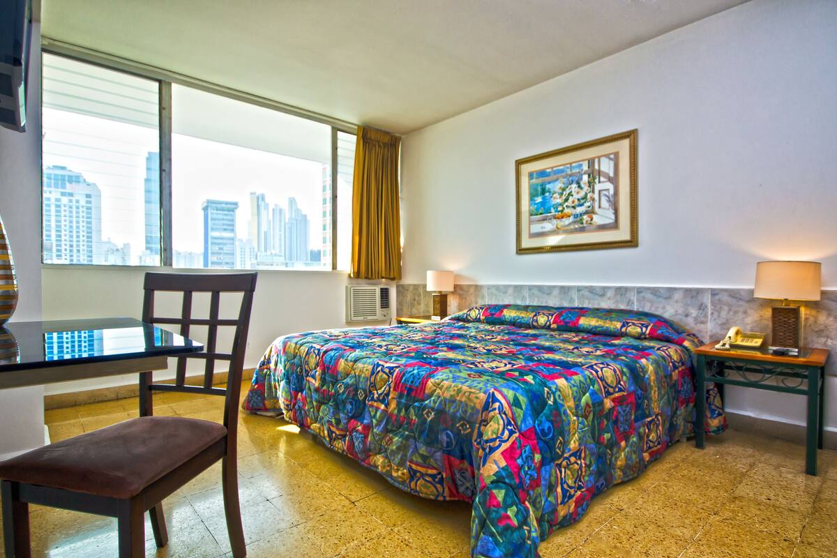 Una habitación de hotel con una cama grande, un escritorio con una silla, una ventana con vista a la ciudad y un cuadro en la pared.