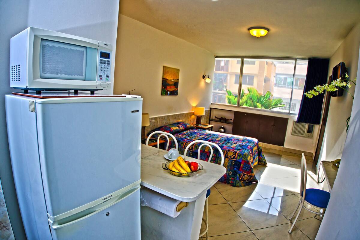 Habitación de hotel compacta con kitchenette, una cama con colcha de colores, una pequeña mesa con fruta y una ventana con vistas a edificios.