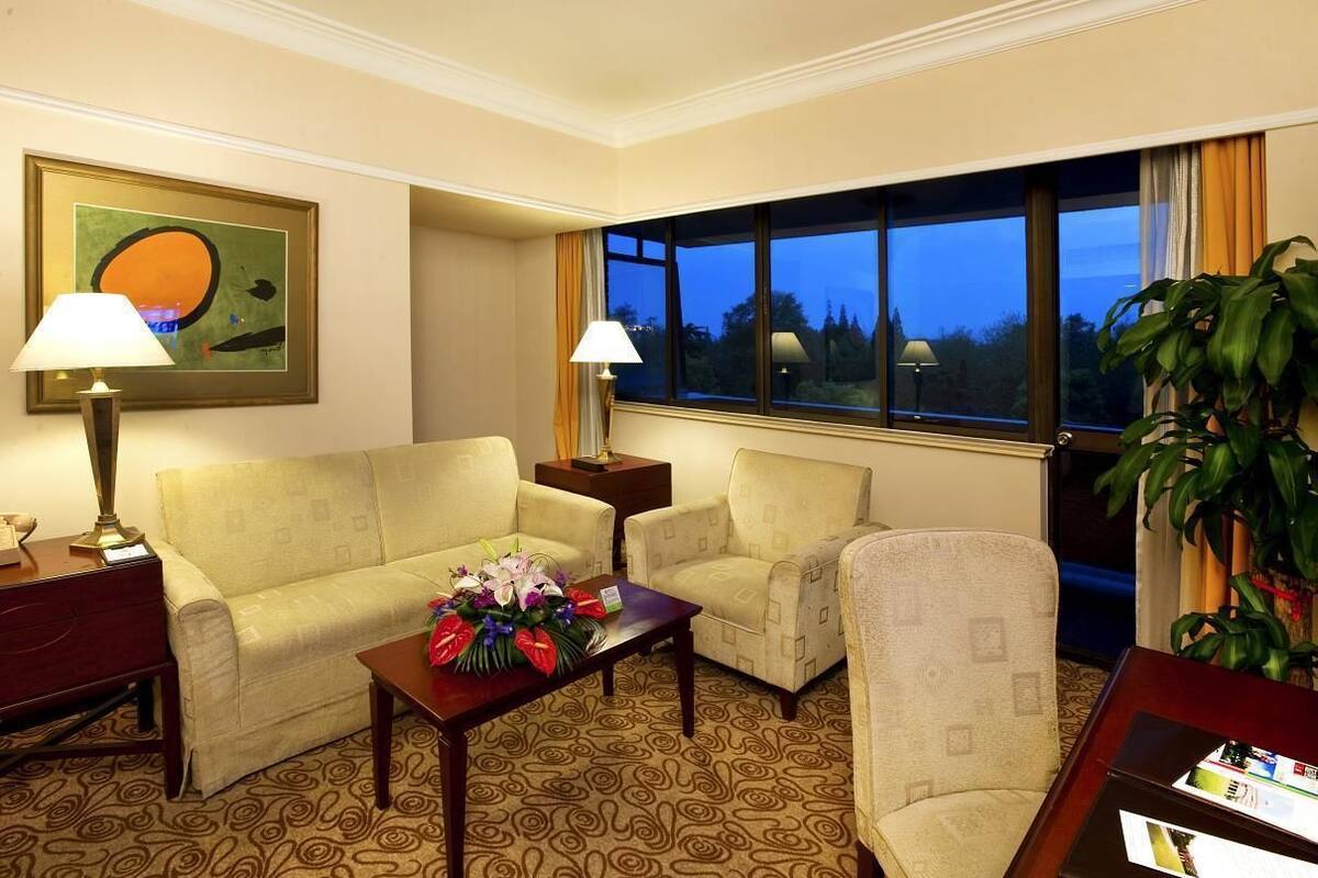 Habitación de hotel con sofá, dos sillones, una mesa de centro con flores, una lámpara, un cuadro y un ventanal con vistas a árboles.