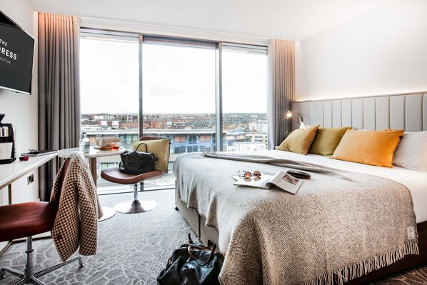 A hotel room with a large bed, a desk, a chair, and a view of the city through large windows.