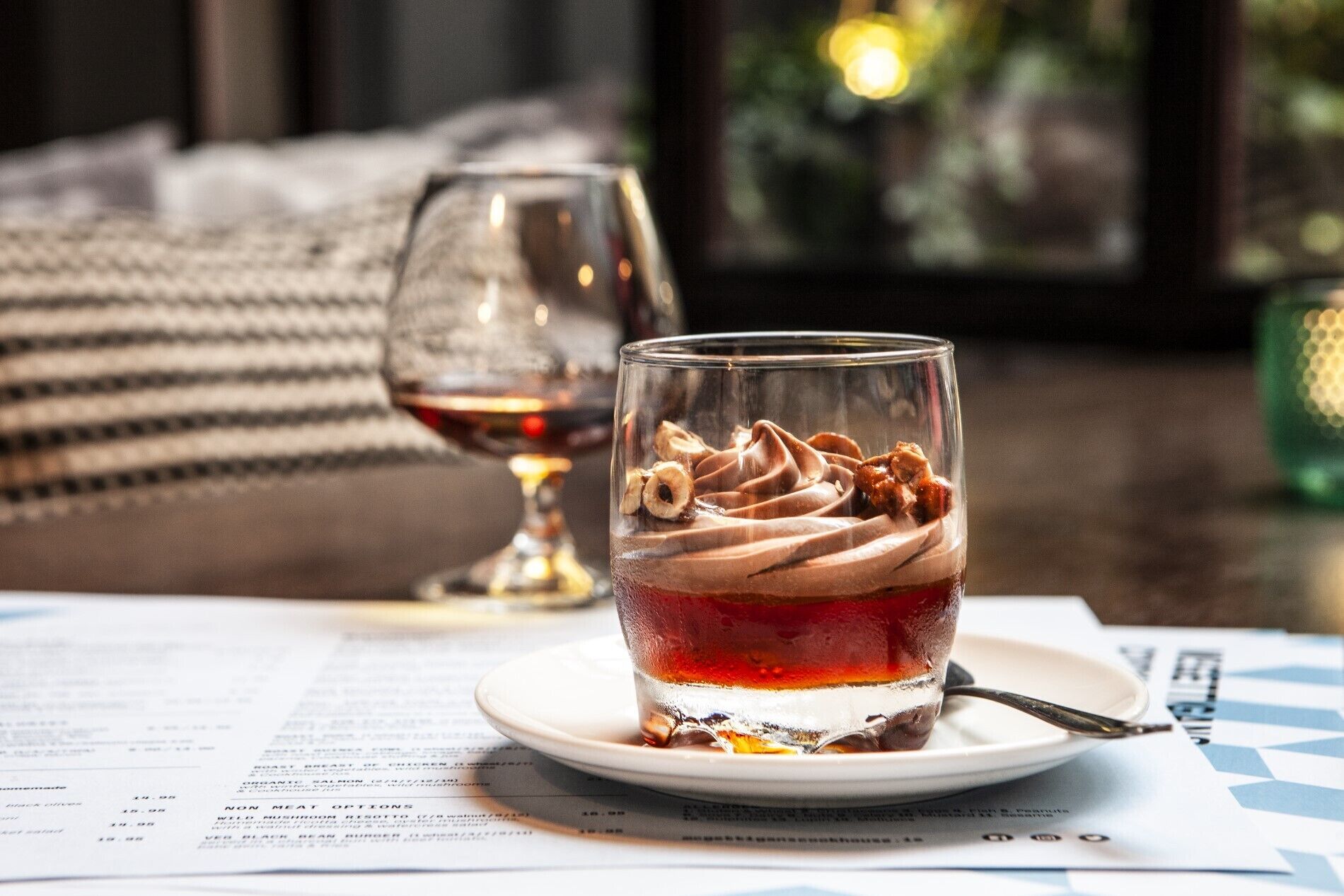 Un vaso de postre con mousse de chocolate y nueces sobre un plato blanco, una copa de vino y un menú en una mesa.
