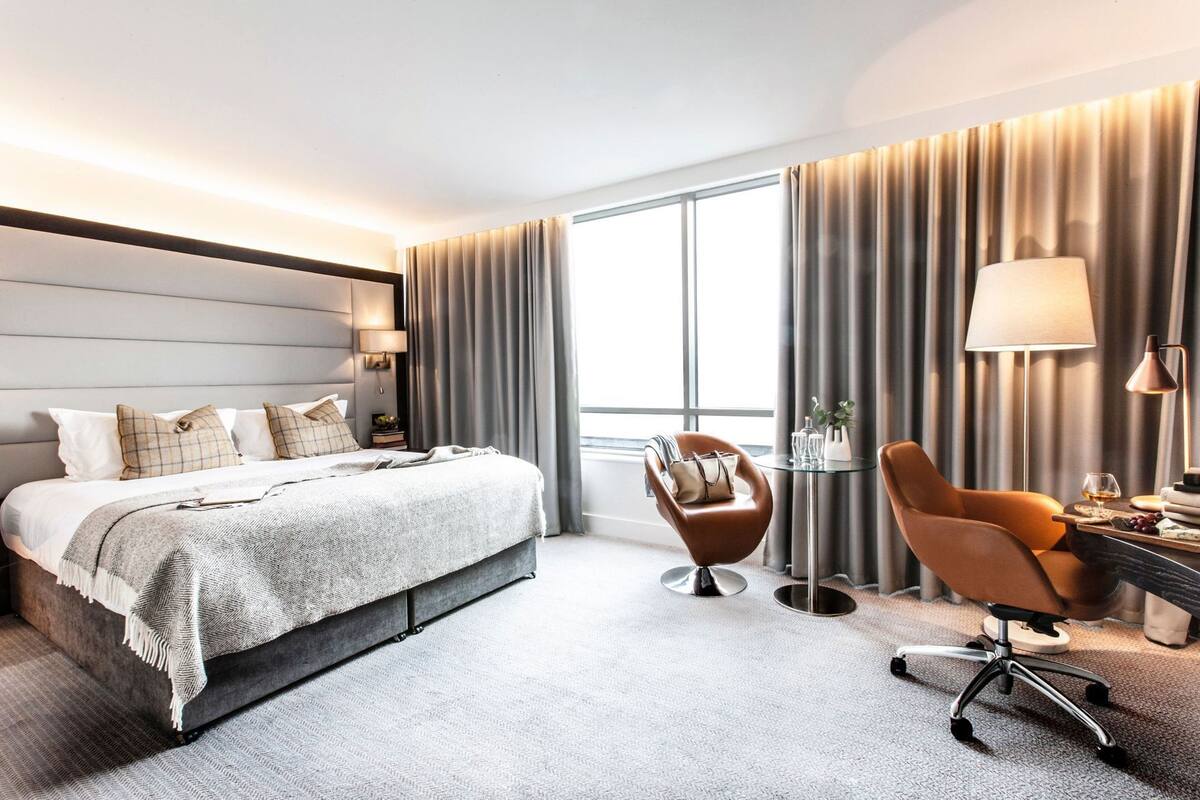 Camera d'hotel moderna con un letto grande, una zona salotto con una poltrona, una scrivania con una lampada e vista sull'esterno.