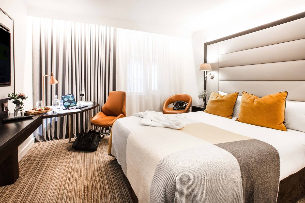 Una camera d'albergo con un letto, una scrivania, una poltrona arancione, una televisione e una finestra con tende.