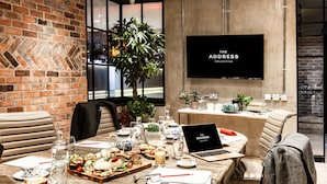 Una sala de reuniones moderna con pared de ladrillo, una pantalla grande mostrando 'THE ADDRESS COLLECTIVE', y una mesa preparada con comida y bebidas.