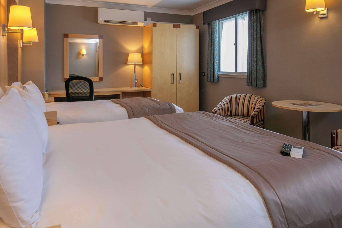 A hotel room with two beds, a desk with a chair, a small table, and a window with curtains.