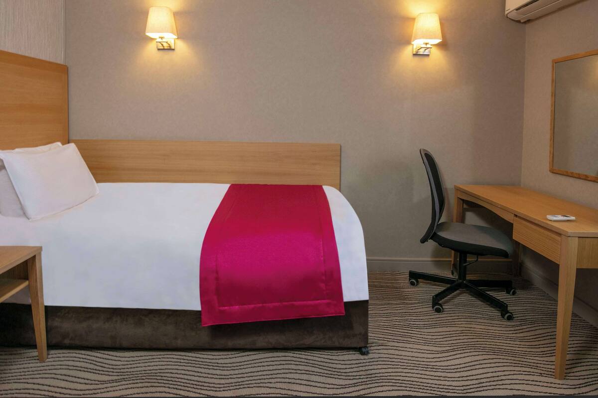 A hotel room with a bed, bedside table, desk, chair, and two wall-mounted lamps.