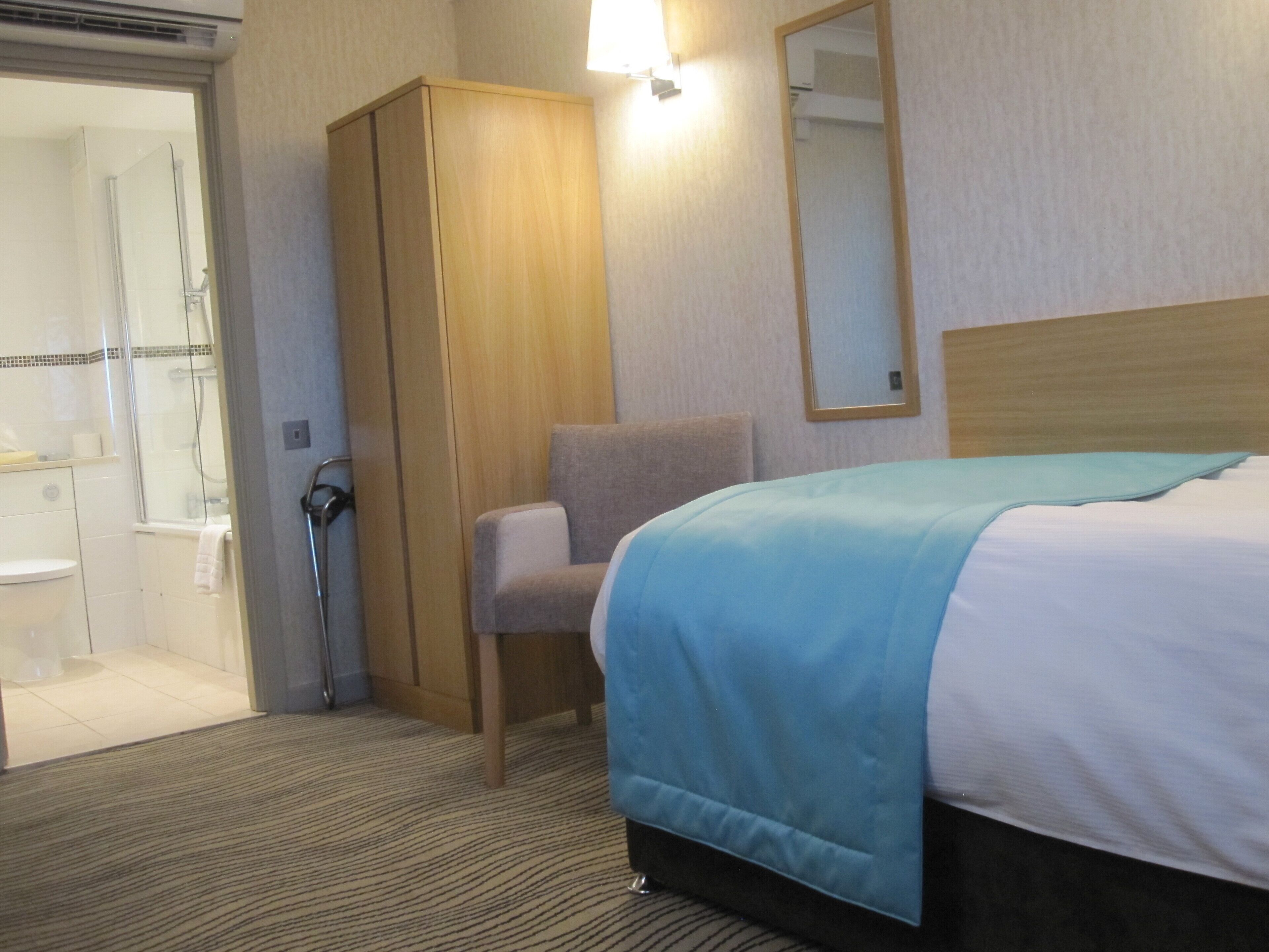 Habitación de hotel con cama, una silla, armario, zona de ducha y espejo.