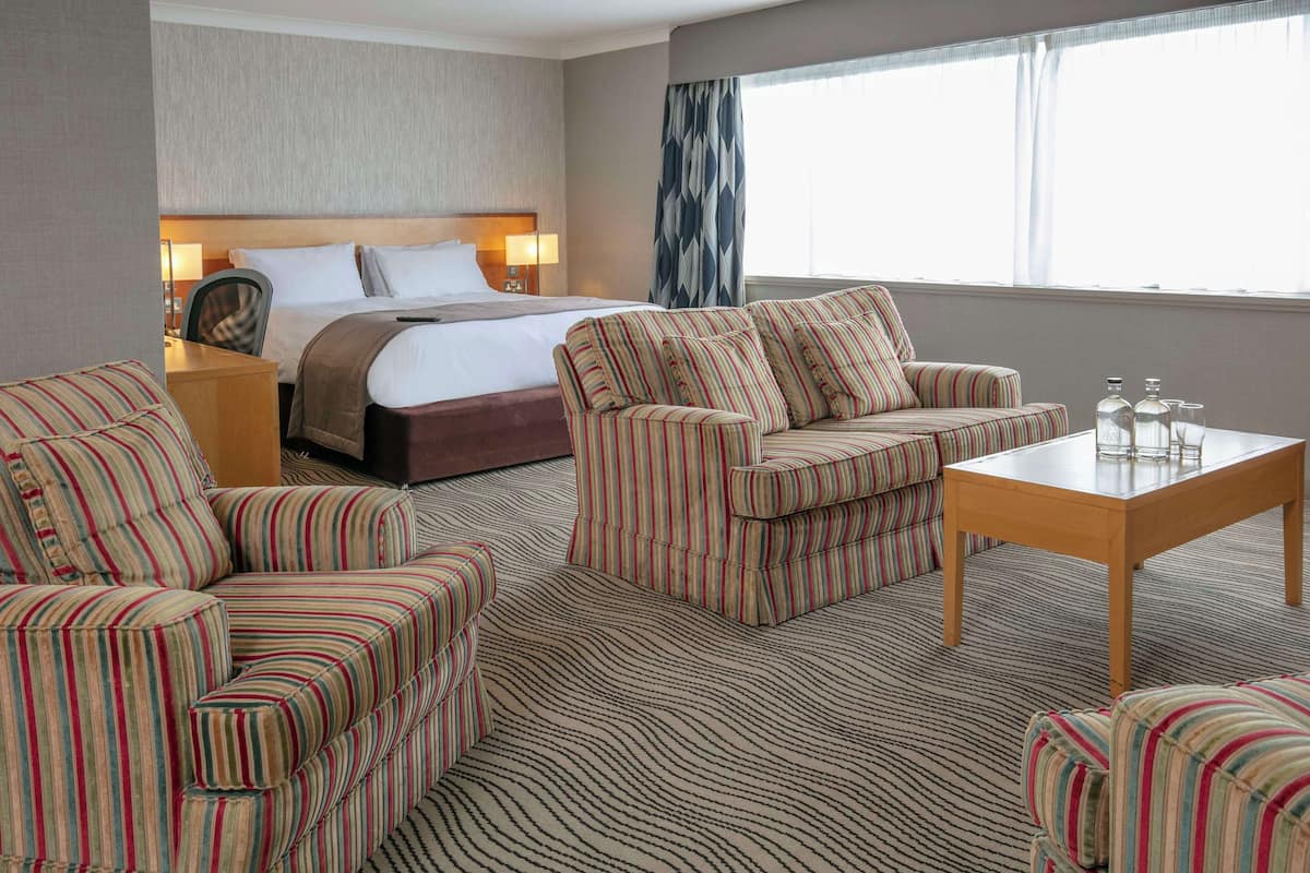 A hotel room with a bed, striped armchairs, a sofa, a wooden table, and a patterned carpet.