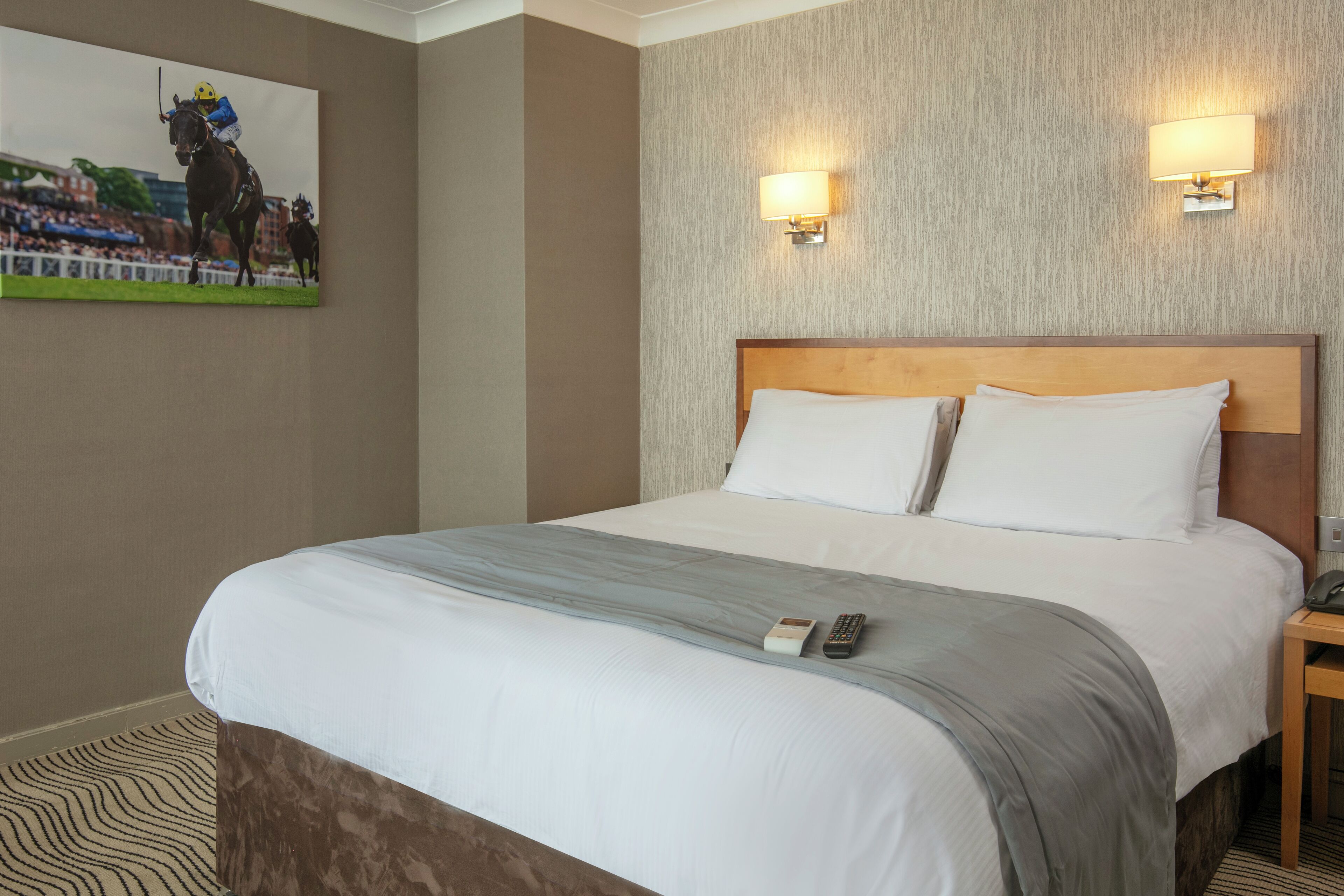 A hotel room with a large bed, two bedside lamps, a mounted picture of a jockey on a horse, and a patterned carpet.