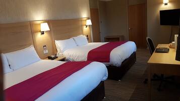 A hotel room with two beds, each with white bedding and a red blanket, a desk with a chair, a television, and a small table with a cup and a book.