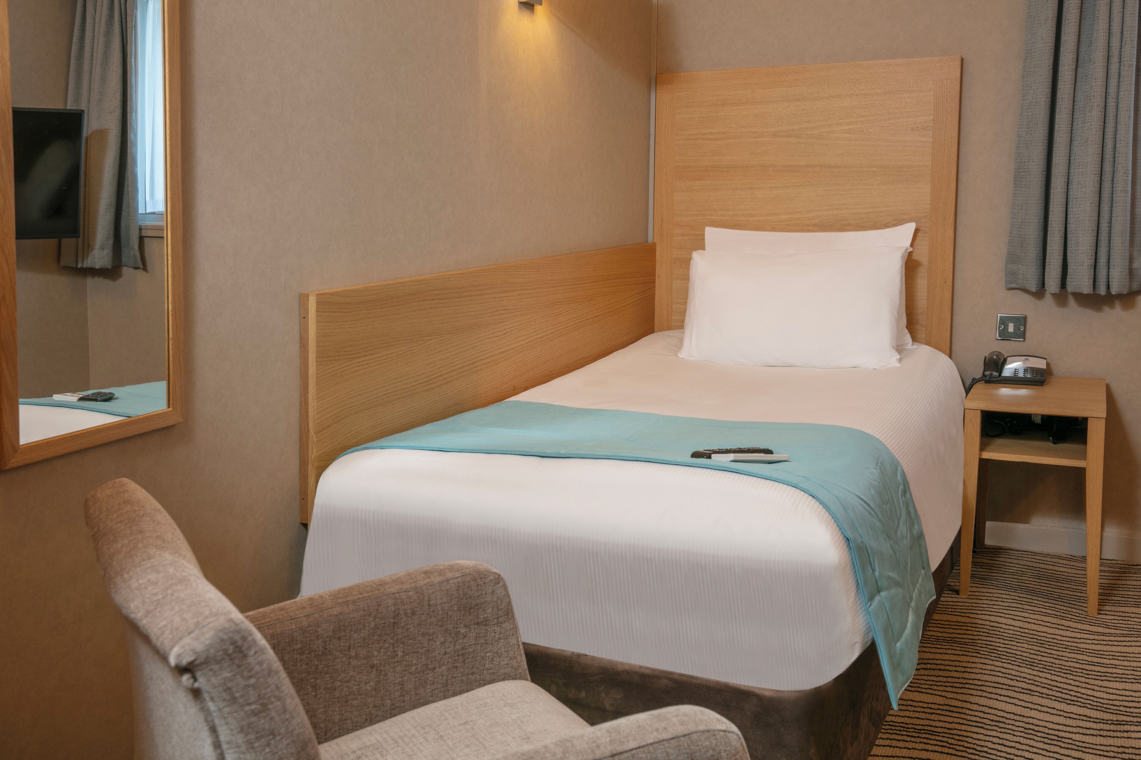 A hotel room with a bed, a chair, a bedside table with a lamp, and a TV mounted on the wall.