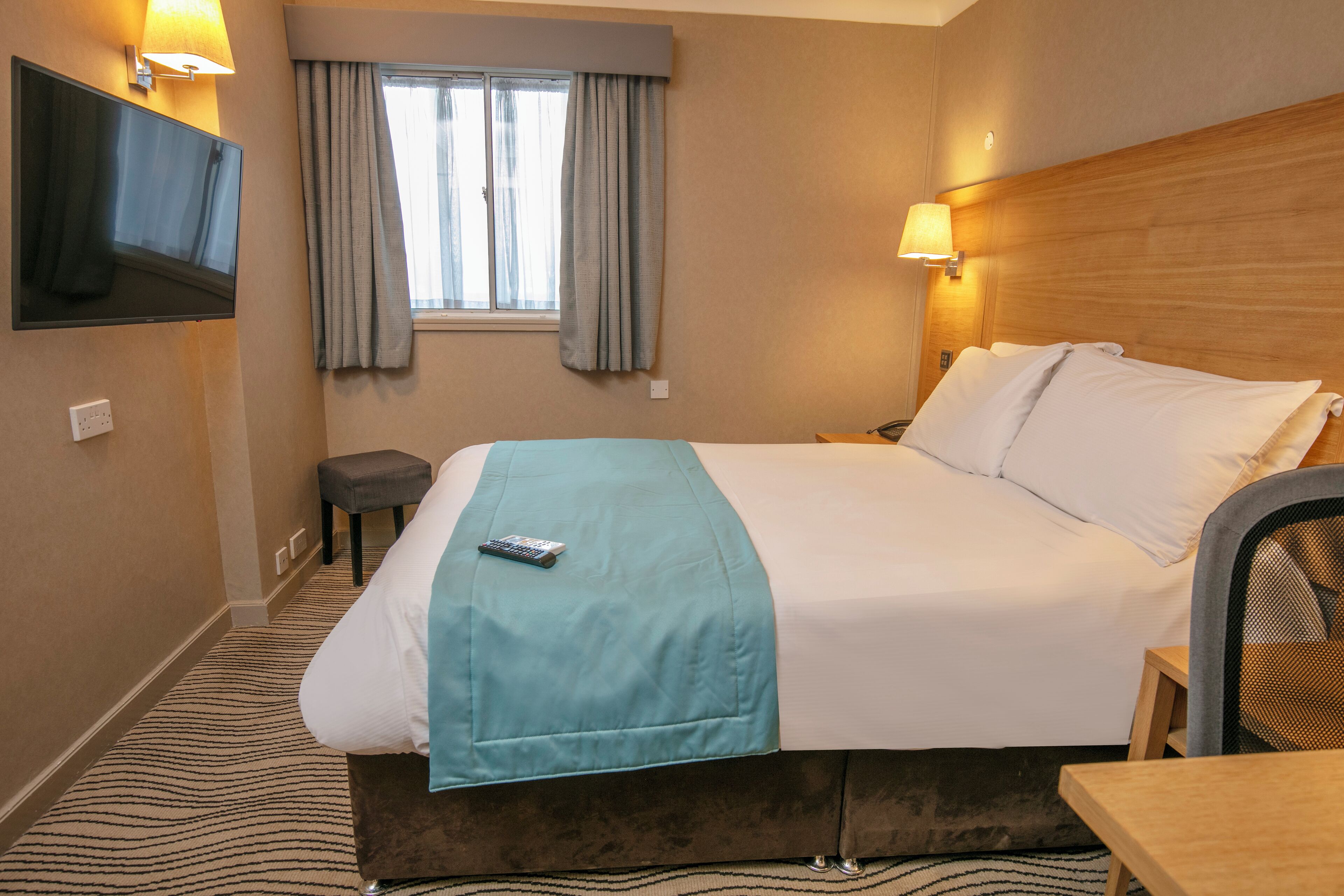 A hotel room with a bed, a TV mounted on the wall, a window with curtains, a bedside table, and a chair.