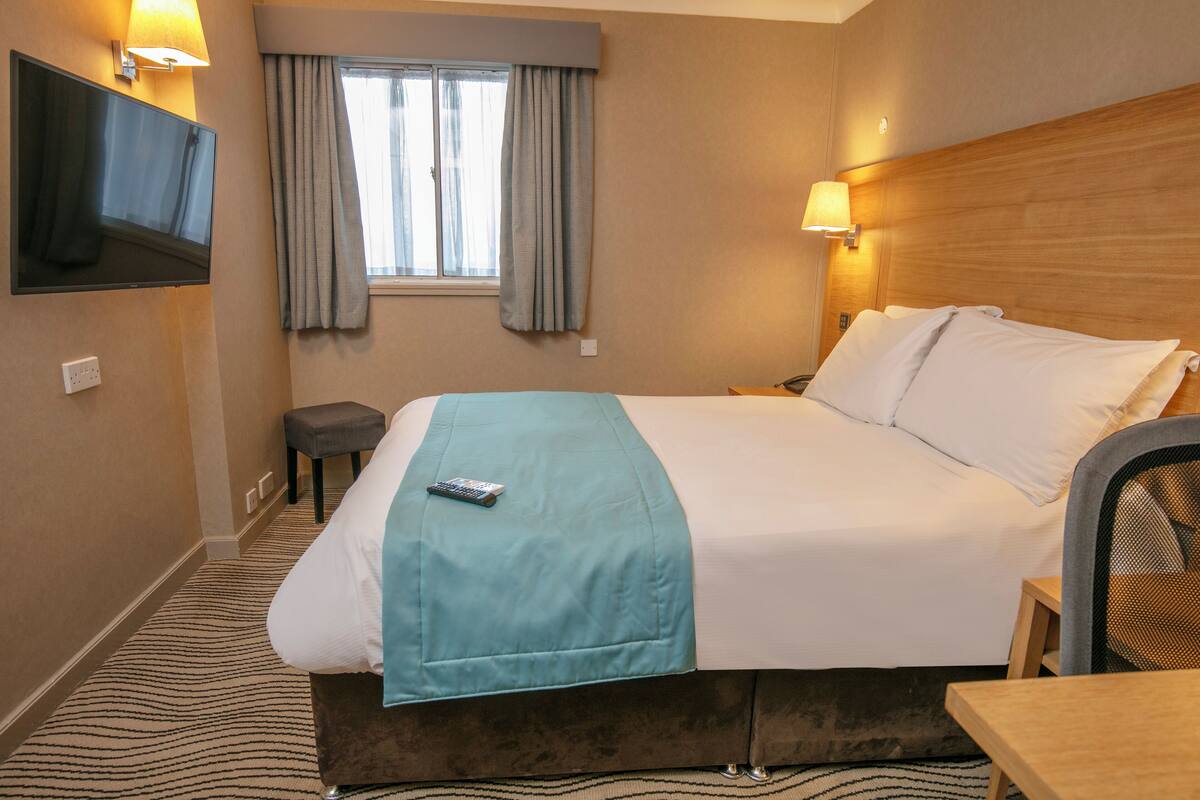 Une chambre d’hôtel comprenant un lit, un téléviseur fixé au mur, une fenêtre avec des rideaux, une table de chevet et une chaise.