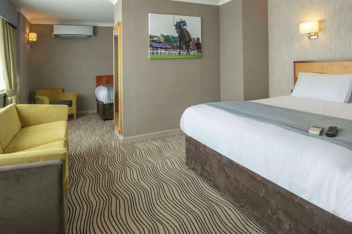 A hotel room with a large bed, a yellow armchair, a small table, and a wall-mounted picture of a horse race.
