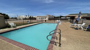 A rectangular pool with a brick edge and a metal safety railing. There are white lounge chairs and a table with umbrellas on the right side. - Gun Barrel Executive Inn (Mabank)