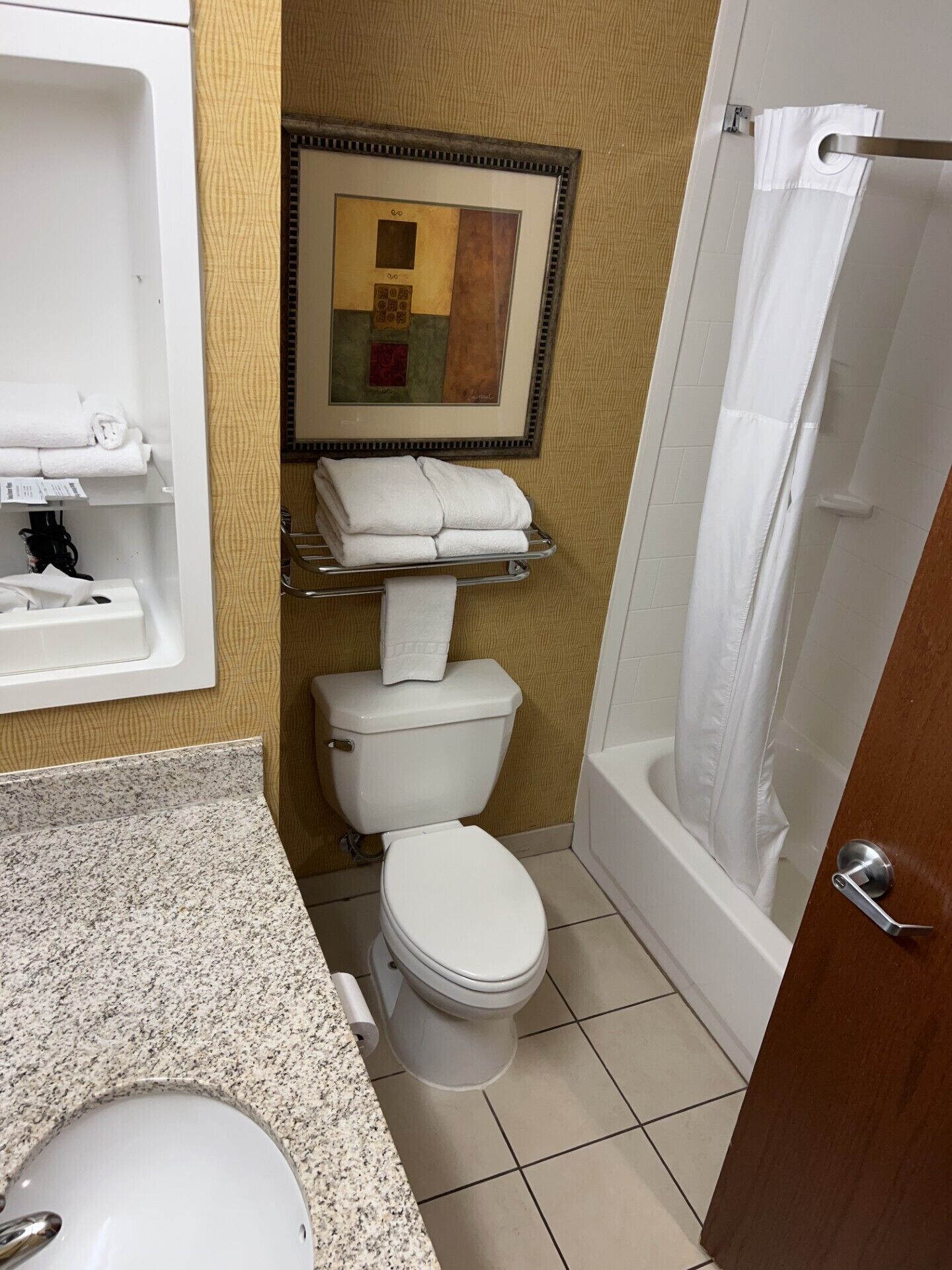 A small bathroom with a toilet, sink, and shower. There is a towel rack with towels and a framed picture on the wall.