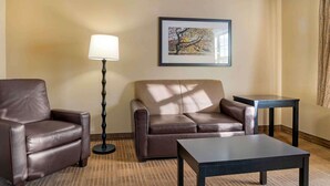A hotel room with a brown leather sofa, a small table, a floor lamp, and a framed picture on the wall. - Extended Stay America Select Suites Detroit Farmington Hills (Farmington Hills)