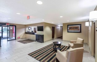 Un hall de hotel con mostrador de recepción, un reloj, un letrero que dice 'BIENVENIDOS', perchero y una zona de estar con sillas y una mesita.