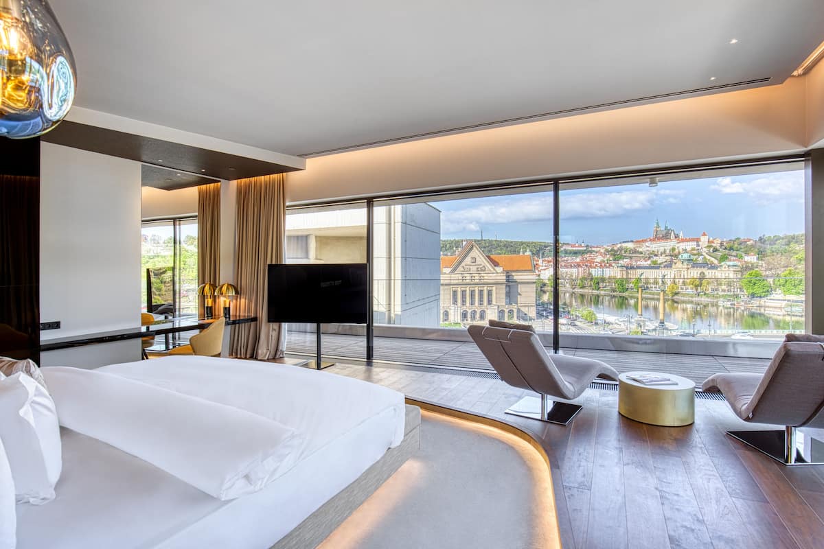 Quarto de hotel moderno com uma cama grande, uma escrivaninha e uma cadeira. Há uma televisão e vista para o horizonte urbano através de amplas janelas.