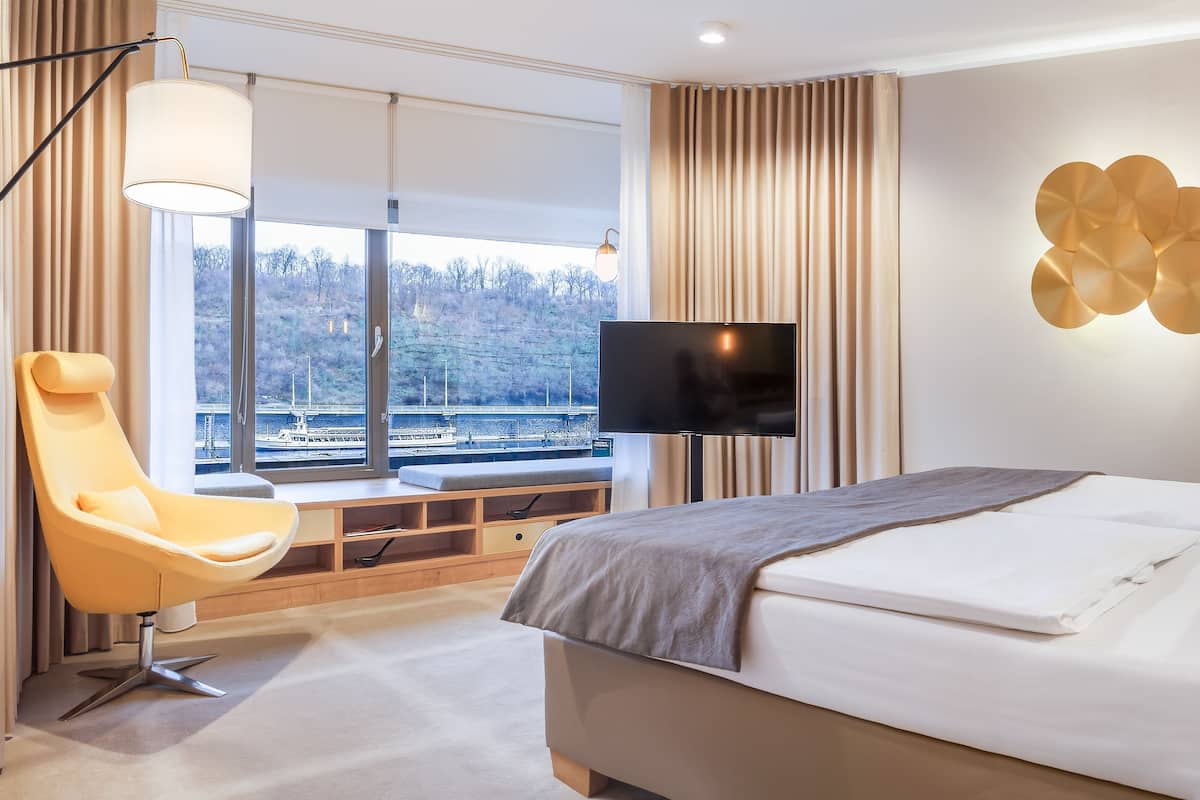 Quarto de hotel moderno com uma cama grande, televisão, uma cadeira e vista para a água.