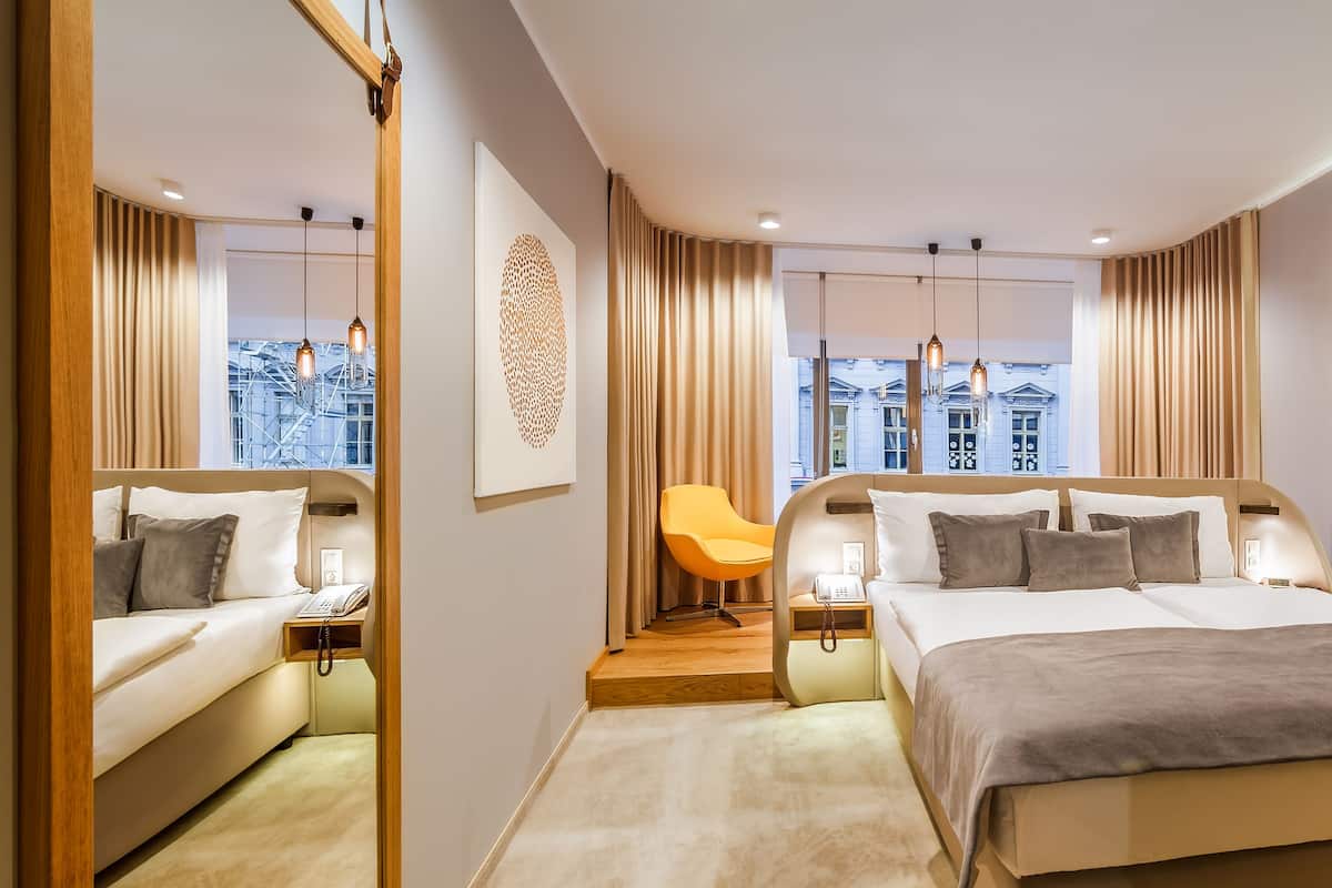 Quarto de hotel moderno com uma cama grande, uma mesa e uma cadeira. Há uma janela com cortinas, um candeeiro fixado na parede e vista para edifícios.