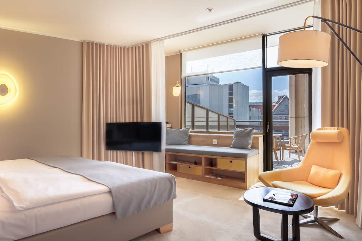 Quarto de hotel moderno com uma cama grande, televisão, poltrona confortável e vista para a cidade.