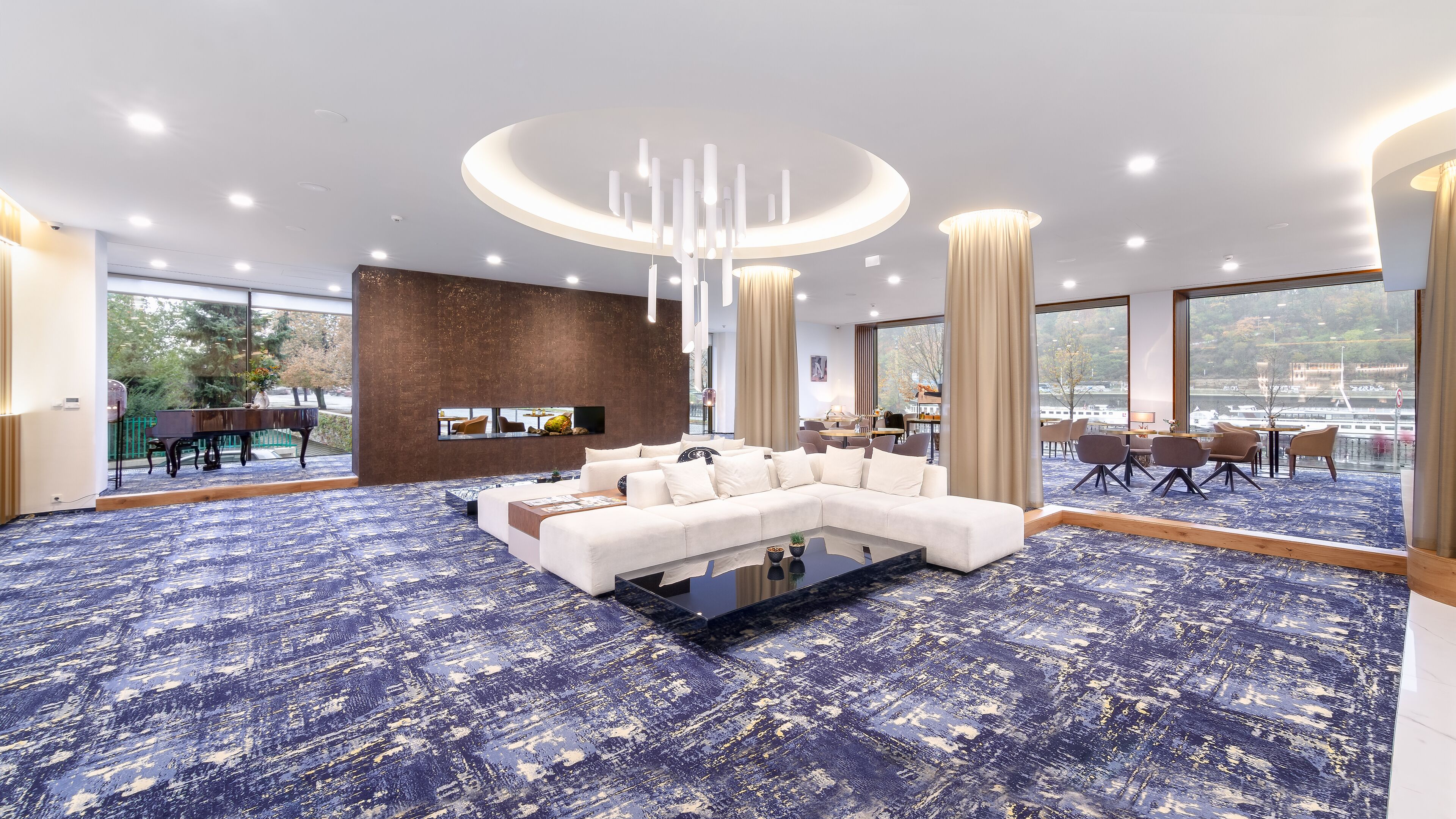 Een moderne hotel lobby met een grote witte modulaire bank, een eetgedeelte en grote ramen met uitzicht op een waterpartij.