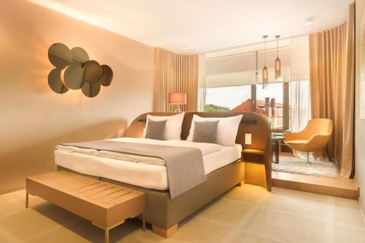 Quarto de hotel moderno com uma cama grande, mesas de cabeceira, um banco, uma cadeira e vista para o exterior.