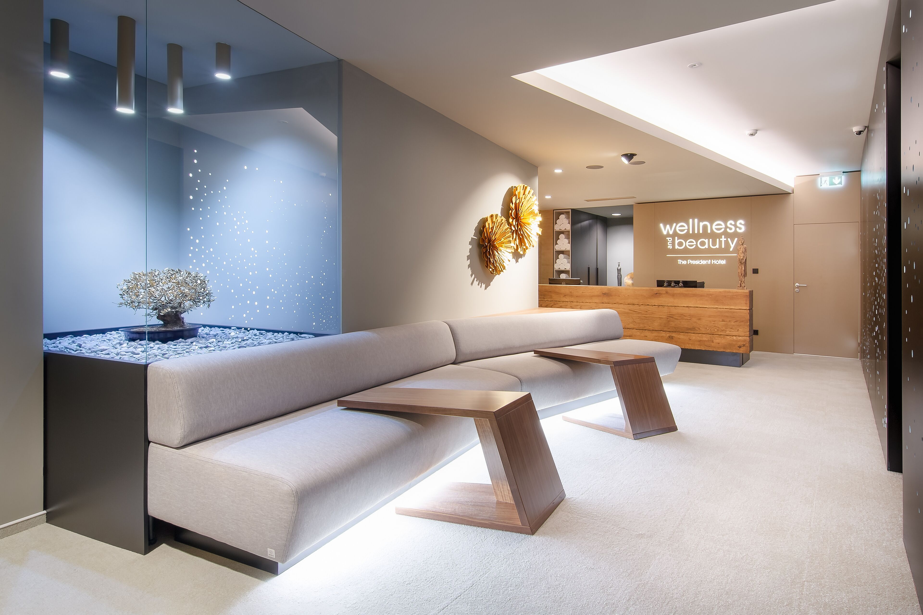 Uma área de recepção moderna, com balcão, espaço de estar com sofá e mesas, e uma parede decorativa com a inscrição 'wellness and beauty'.