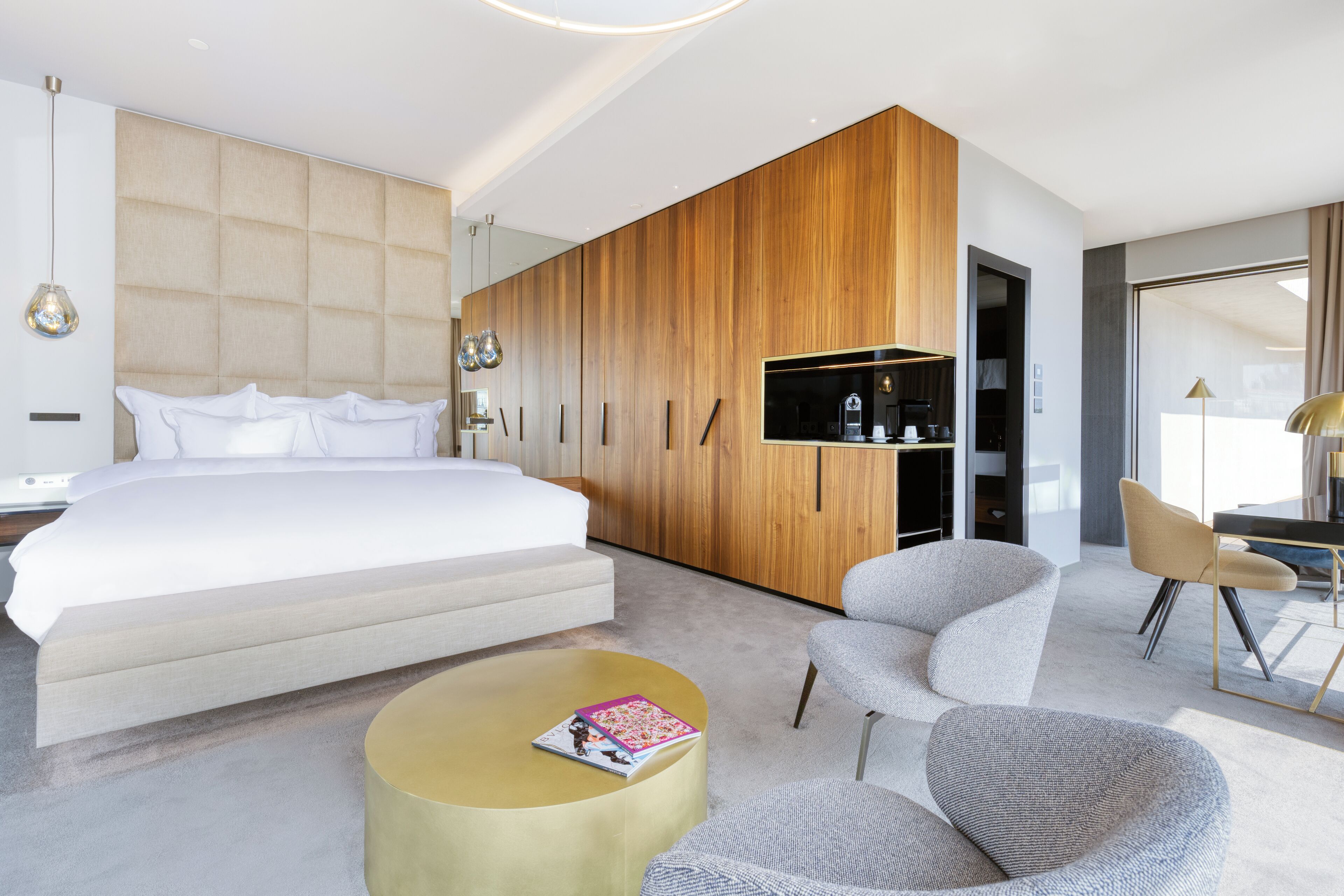 A modern hotel room with a large bed, a wooden wardrobe, a round coffee table, and a seating area with chairs.