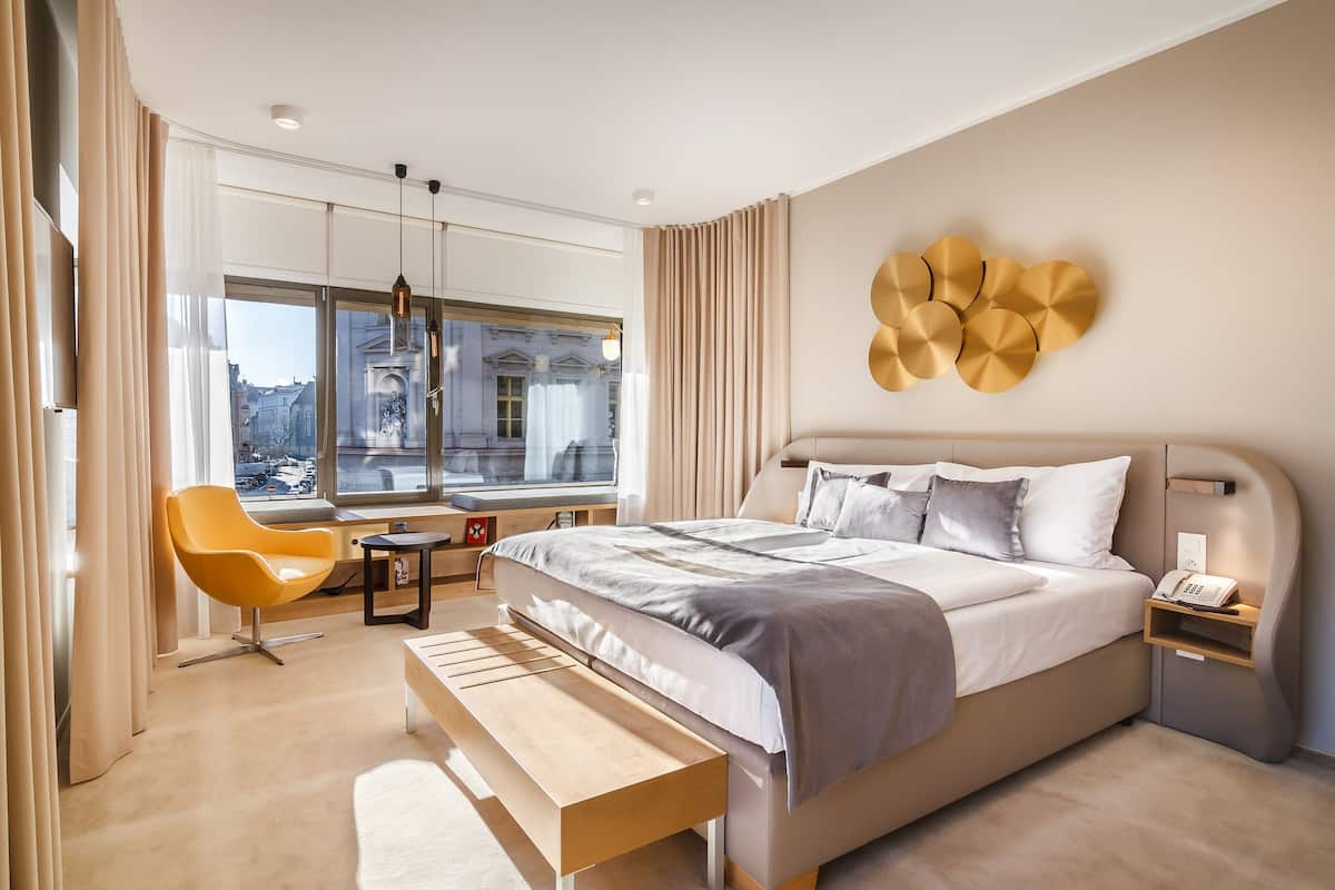 Quarto de hotel moderno com uma cama grande, uma mesinha de cabeceira com telefone, um banco, uma cadeira amarela e vista para a cidade através de amplas janelas.