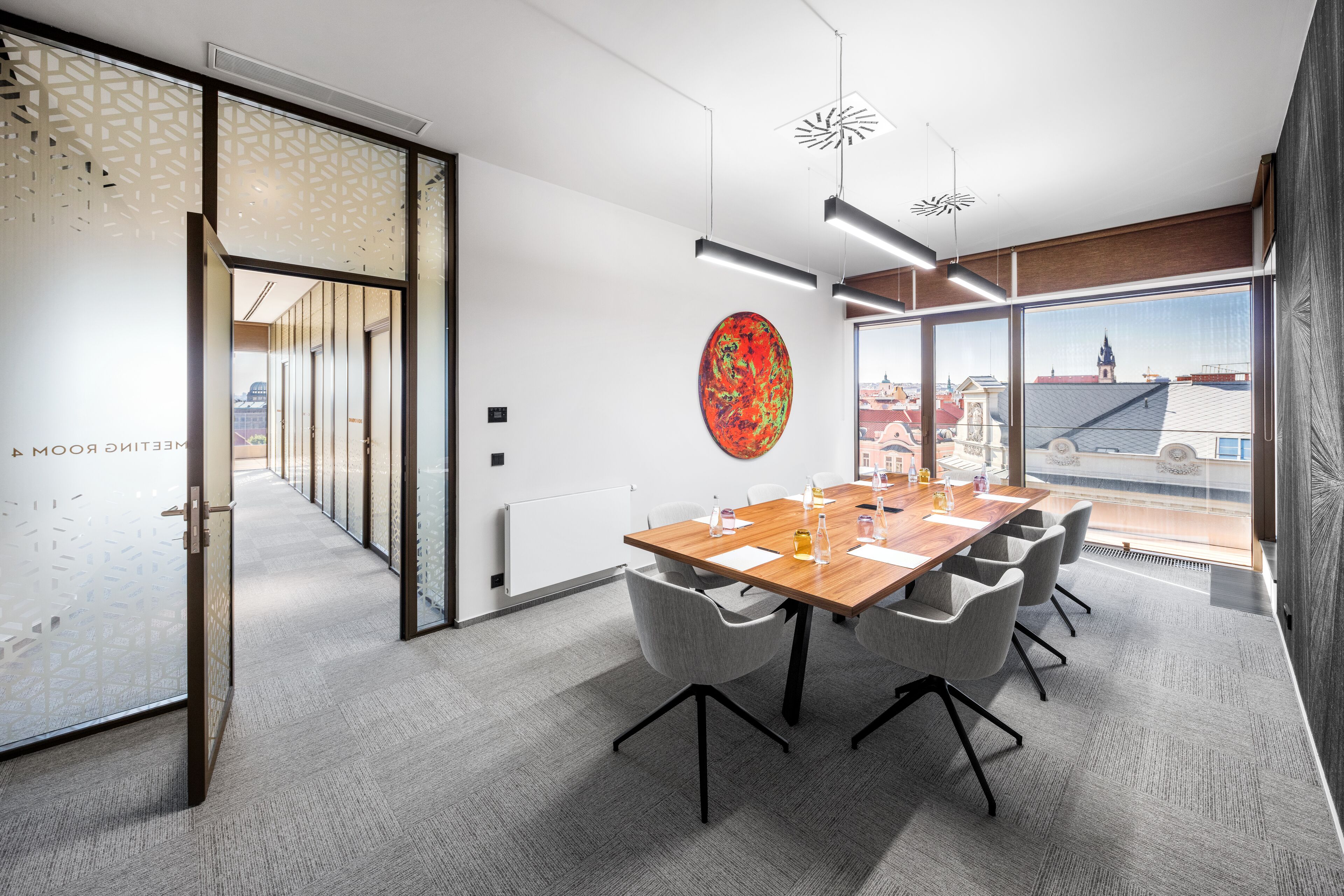 Ein moderner Konferenzraum mit Holztisch, grauen Stühlen, einem großen Fenster mit Blick auf Gebäude und einem Wanddekor.
